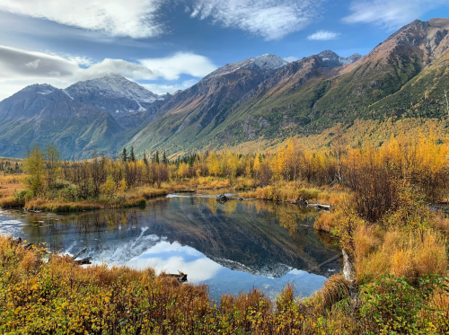 A serene landscape featuring a calm pond reflecting mountains and autumn foliage under a blue sky with clouds.